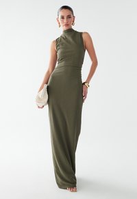 Oliven grøn, ribbet maxikjole med høj halsudskæring, tætsiddende liv og flagrende skørt. Model som holder en struktureret cremefarvet clutch. Enkle smykkedesigns.