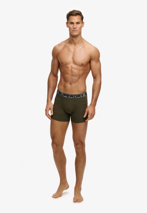 Homme musclé debout pieds nus portant un boxer vert foncé avec une ceinture grise portant la marque Superdry sur un fond blanc uni.