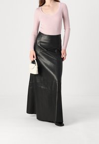 Haut rose à manches longues côtelé associé à une jupe maxi en cuir noir taille haute, tenant un petit sac à main blanc. Texture lisse, design ajusté.