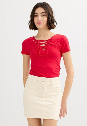 Joven mujer con cabello oscuro y ondulado, que lleva una blusa roja con cordones y mangas cortas y una falda crema de talle alto, de pie frente a un fondo liso.