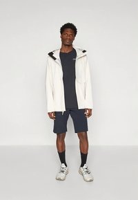 Bela dežna jakna s črnimi poudarki, mornarsko modra majica, mornarsko modre kratke hlače, črne nogavice in svetlo sive športne superge. Model stoji pred enobarvnim ozadjem.