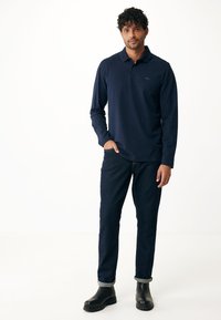 Polo a maniche lunghe blu navy con colletto, abbinato a jeans di denim scuro e stivali alla caviglia neri, caratterizzato da una vestibilità slim e maniche arrotolate.
