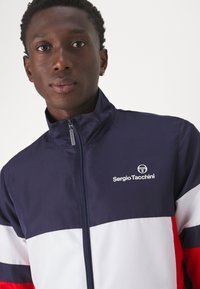 Navy zip-up jacket featuring white and red horizontal stripes, made of smooth, lightweight material. Logo displayed on the upper left.