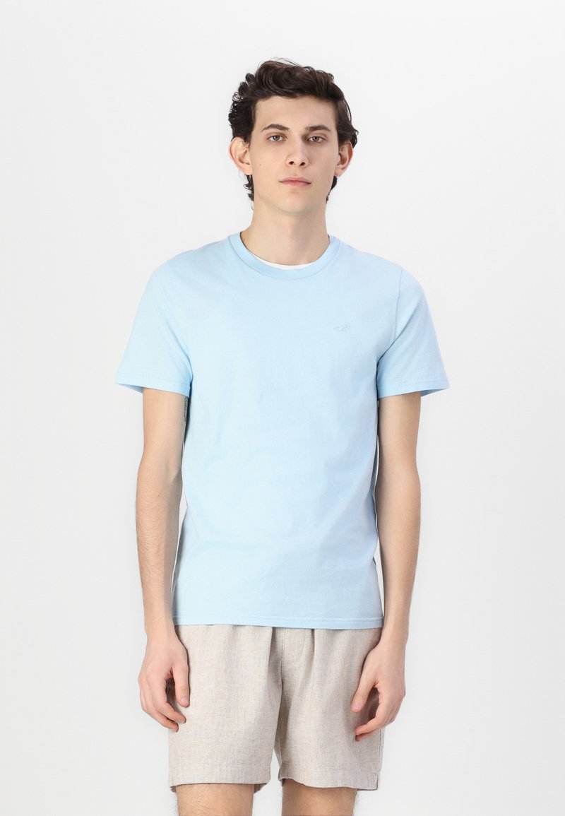 Ανοιχτού μπλε βαμβακερό μπλουζάκι με κοντά μανίκια και στρογγυλή λαιμόκοψη. Διαθέτει ένα διακριτικό λογότυπο στο στήθος και συνδυάζεται με ανοιχτόχρωμο σορτσάκι.