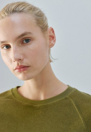 Giovane donna con i capelli biondi raccolti, che indossa un maglione verde oliva, che guarda leggermente di lato su uno sfondo semplice.