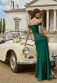 Žalia off-the-shoulder suknelė su prigludusia viršutine dalimi, krentanti sijoninė dalis; modelis remiasi į vintage grietinėlės spalvos automobilį, papuoštą baltomis gėlėmis.
