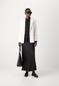 Weißer maßgeschneiderter Blazer über einem schwarzen Hemd mit Knöpfen und einem schwarzen Maxirock, kombiniert mit schwarzen Schuhen und einer schwarzen Tragetasche.
