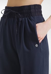 Granatowe spodnie wykonane z miękkiego materiału, wyposażone w elastyczny pas ze sznurkiem, boczne kieszenie oraz małe logo na dolnym boku.