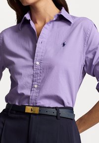 Camisa de algodón color lila claro con botones, que presenta un pequeño logo azul marino, combinada con un cinturón de cuero azul oscuro y pantalones oscuros.