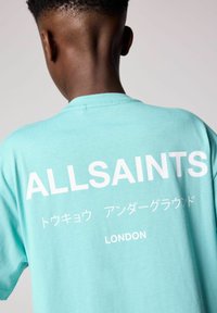 Turkos bomullströja med vit text som lyder "ALLSAINTS," japansk skrift och "LONDON," som visar en avslappnad passform och mjuk textur.