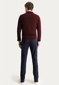 Bordeauxroter Strickpullover mit einem gerippten Saum, kombiniert mit navyblauen Hosen und weißen Sneakers. Von hinten gezeigt, mit einem einfachen Design.
