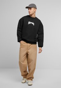 Zwarte sweatshirt met oversized pasvorm, witte grafische tekst, gecombineerd met tan wijdvallende broek en witte sneakers. Grijze pet maakt de look af.