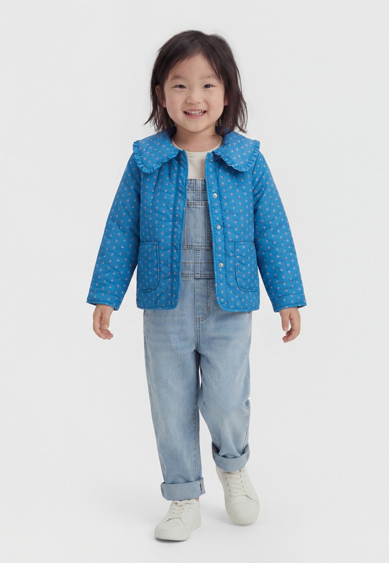 Χαμογελαστό παιδί που φοράει μπλε ζακέτα με σχέδιο πάνω από τζιν φορμάκι και λευκά αθλητικά, στέκεται μπροστά σε απλό λευκό φόντο.