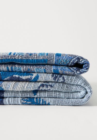 Tissu tissé plié avec des motifs géométriques bleus, blancs et gris empilés sur une surface blanche contre un fond uni.