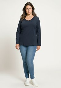 Námořnická dlouhá vesta s výstřihem do V v kombinaci s modrými slim-fit džíny a bílými teniskami. Tato vesta má uvolněný střih a jemnou texturu.