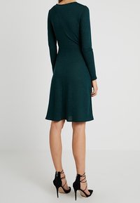 Tmavě zelené šaty s dlouhým rukávem vyrobené z texturované tkaniny, rozšiřující se u hemu. V páru s černými sandály na vysokém podpatku se šněrováním.