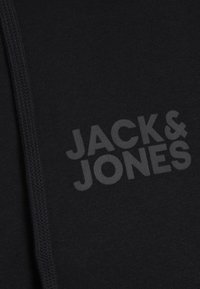 Tissu noir avec un logo "JACK & JONES" légèrement en relief imprimé en gris foncé près d'un cordon épais sur le côté gauche.