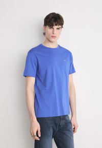 Blaues Baumwoll-T-Shirt mit rundem Ausschnitt und kurzen Ärmeln, versehen mit einem kleinen Logo-Detail auf der linken Brust. Kombiniert mit dunklen Jeans.