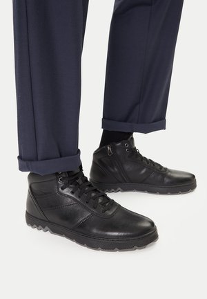 Czarne, wysokie buty ze skóry z teksturowaną powierzchnią, sznurowane, z gumową podeszwą. Noszone z granatowymi spodniami, które są podwinięte na dole.