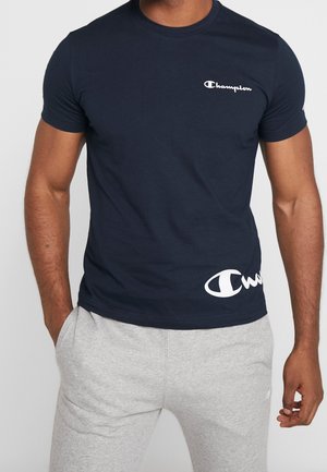 Mann trägt ein marineblaues Champion-T-Shirt mit weißem Logotext, kombiniert mit hellgrauen Jogginghosen, steht vor einem schlichten Hintergrund.