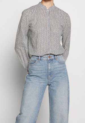 Persona con blusa blanca de manga larga con estampado negro metida en unos vaqueros de tiro alto color azul claro, de pie con las manos detrás de la espalda.