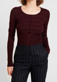 Haut bordeaux à manches longues avec un détail côtelé texturé, col rond et silhouette ajustée, associé à un pantalon noir à fines rayures.