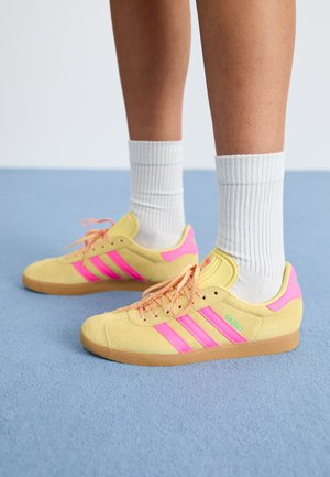 Baskets en daim jaunes avec des accents roses, trois bandes et une marque. Semelle en caoutchouc et lacets roses, portées avec des chaussettes blanches à rayures.