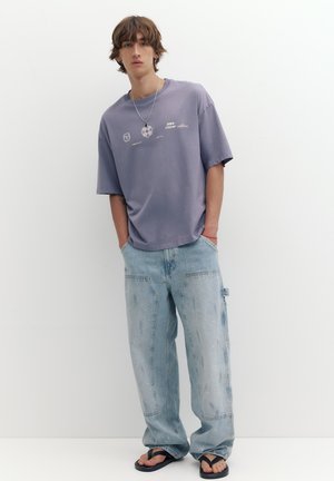 Jeune homme aux cheveux bruns ébouriffés porte un t-shirt graphique lavande ample, un jean large bleu clair, des sandales noires et un collier en argent.