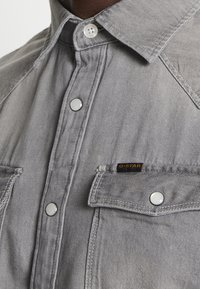 Camicia in denim grigia con colletto a punta, bottoni a pressione, due tasche sul petto e un piccolo logo G-Star sulla tasca.