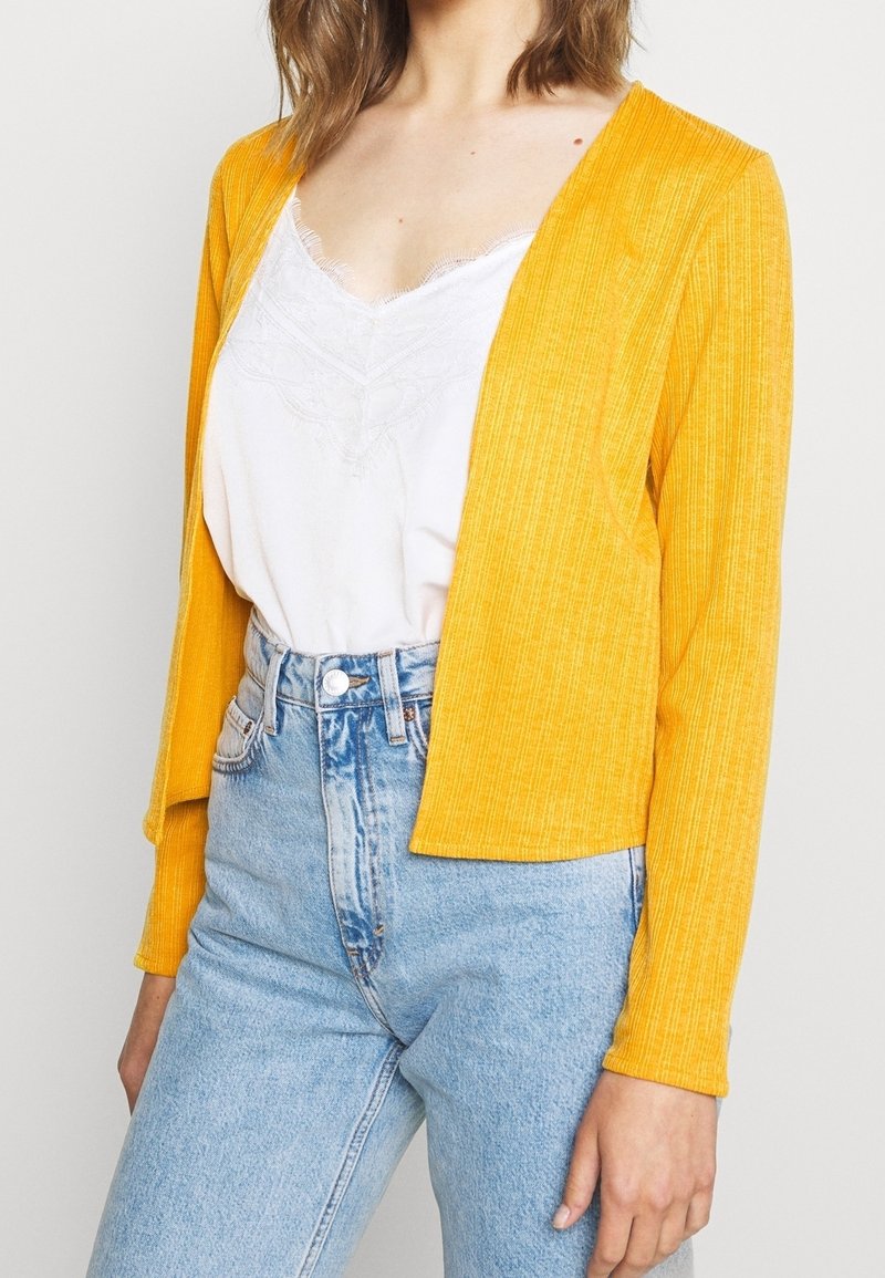 Femme portant un jean taille haute bleu clair, un top blanc bordé de dentelle et un cardigan jaune côtelé ouvert devant, debout devant un fond uni.