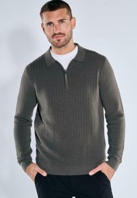 Dunkelgrüner, strukturierter Pullover mit Reißverschlusskragen und kontrastierendem, einfarbigem Kragen. Lange Ärmel mit Gerippteffekten an den Bündchen und dem Saum.