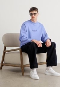 Világoskék pulóver "BARBOUR" logóval, fekete nadrág és fehér cipő. A modell napszemüveget visel, egy fa keretes székben ül.
