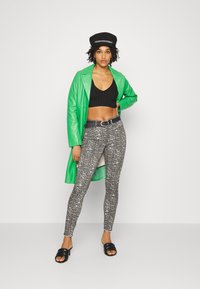 Casaco longo de couro verde, top cropped preto canelado, leggings cinzentas com padrão de cobra, cinto preto e sandálias pretas de slip-on. Texturas suaves e design ajustado.
