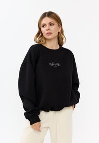 Felpa nera oversize realizzata in morbido tessuto, con collo rotondo e un logo decorativo argentato sul davanti. Vestibilità comoda e maniche lunghe.