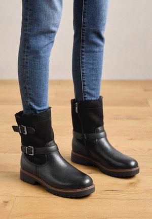 Schwarze Knöchelstiefel aus Leder und Textil, mit doppelten Schnallen, einem seitlichen Reißverschluss und einer strukturierten Sohle. Kombiniert mit eng anliegenden blauen Jeans.