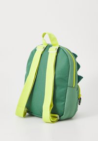 Green backpack with yellow straps, textured material, dinosaur-inspired spikes on the back, and a front zip pocket. Compact design.