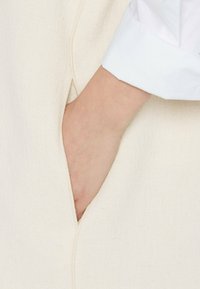 Hand in die Seiten Tasche eines cremefarbenen Stoffs gesteckt, mit einem weißen Hemdmanschette sichtbar am Handgelenk.