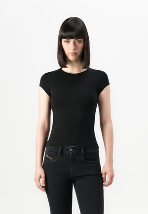Woman with straight dark hair wearing a fitted black short-sleeve top and black jeans, standing against a plain white background.