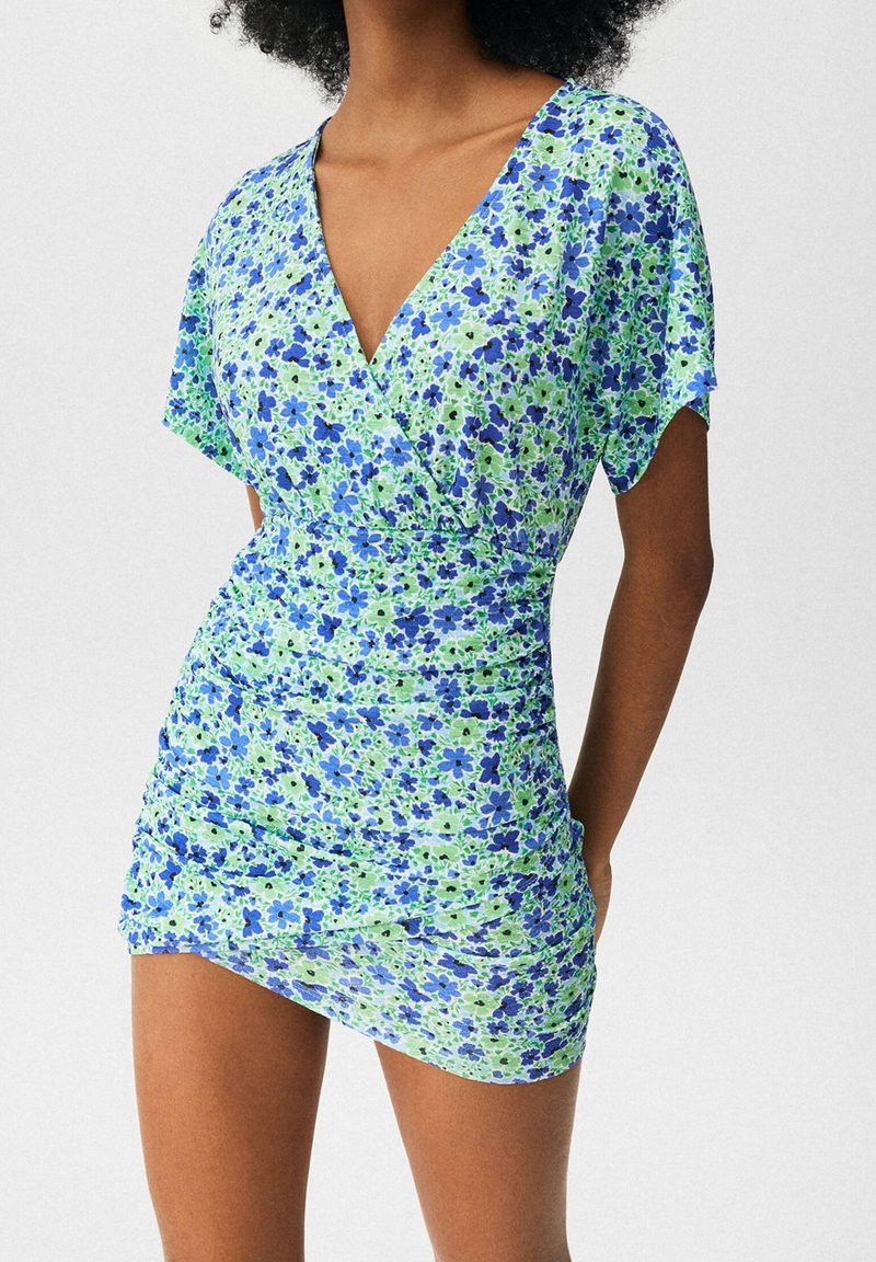 Robe fleurie verte et bleue, avec un design portefeuille, des manches courtes et une texture froncée. Fabriquée en tissu léger et extensible.