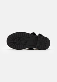 Suela de zapato de goma negra con patrones de tracción, logotipos y orificios de drenaje. Forma plana con bordes redondeados y una textura general lisa.