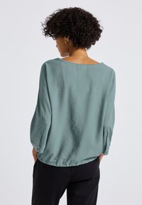 Blusa verde chiaro, morbida e ampia, con maniche a tre quarti, scollatura ampia e finitura texturizzata. Sul retro presenta un dettaglio di cucitura verticale.