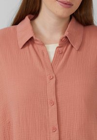 Chemise à boutons de couleur pêche en tissu texturé. Présente un col, une fermeture à boutons sur le devant et une surface légèrement froissée.