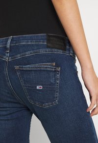 Vaqueros de mezclilla azul oscuro con un diseño ajustado, que cuenta con una etiqueta de cuero negro con 'TOMMY JEANS' y un pequeño logotipo en el bolsillo trasero.