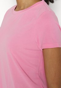 Gros plan d'une personne portant un t-shirt rose clair uni à manches courtes avec un col rond et des coutures visibles.
