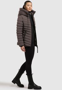 Giovane donna in una giacca imbottita marrone, leggings neri e stivali neri in piedi davanti a uno sfondo grigio uniforme.