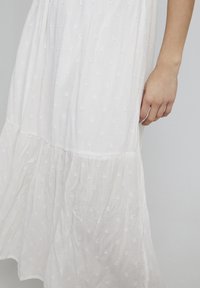 Biała sukienka warstwowa wykonana z teksturowanej tkaniny z delikatnym kropeczkowym wzorem; posiada zwiewny dół i krótkie rękawy.