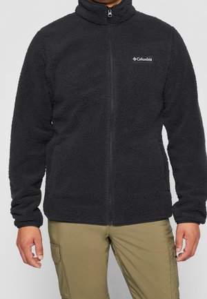 Mann trägt schwarze Fleecejacke mit durchgehendem Reißverschluss und Columbia-Logo auf der Brust sowie beige Cargohosen, steht vor einfarbigem Hintergrund.