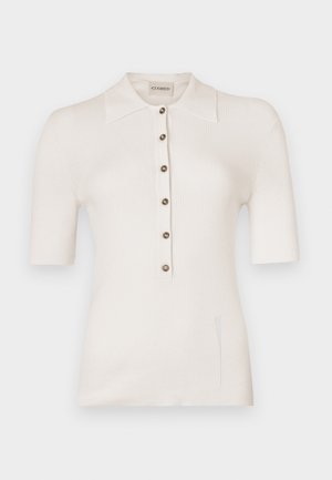 Bílá žebrovaná pletená polo košile s krátkými rukávy, zapínáním na knoflíky vpředu a bočními rozparky. Má klasický límec a kulaté knoflíky.