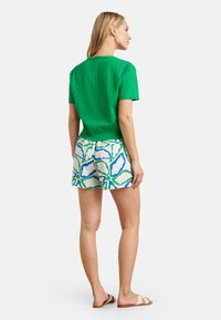 Grünes Kurzarmshirt mit strukturiertem Muster, kombiniert mit weißen Shorts mit blauem und grünem abstraktem Design und beigen Rutschpantoletten.