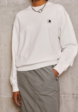 Persoon draagt een wit Carhartt sweatshirt en een grijze rok, hand in de zak, met een zilveren ketting met twee hartvormige hangers.